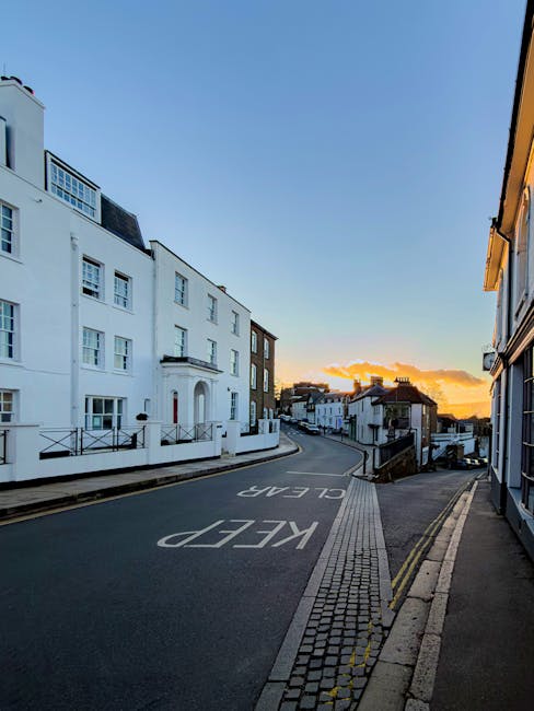 A narrow residential street in Harrow at sunset, featuring white multi-storey buildings with balconies on the left side and a mix of terraced houses on the right. The road is marked with a large white 'KEEP CLEAR' sign painted on the asphalt, and the pavement is made of paving stones along the edges. In the foreground, a parked van with an open rear door is visible, presumably being loaded or unloaded during a home relocation process by Man with Van West Harrow. No furniture or moving boxes are visible in this image, but the scene suggests the logistics of a house removal or furniture transport is underway or planned, with the urban environment providing a typical setting for moving services in the area.