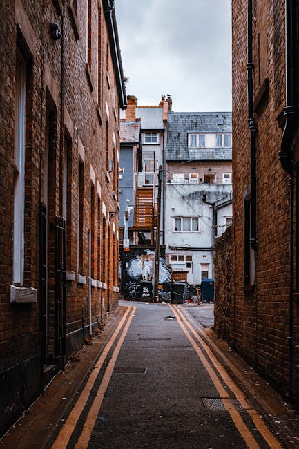 A narrow urban passageway between two brick buildings during daytime, with a double yellow line running along the pavement. In the background, a residential building with a staircase and external fire escape, along with windows and chimneys, are visible. The alley contains several black garbage bins and a large plastic-wrapped item, possibly furniture or boxes, indicating a home relocation or moving process. Man with Van West Harrow's removal team may be involved in the loading or unloading activity, with the scene portraying a typical context for furniture transport and packing during a house move in West Harrow, Harrow.