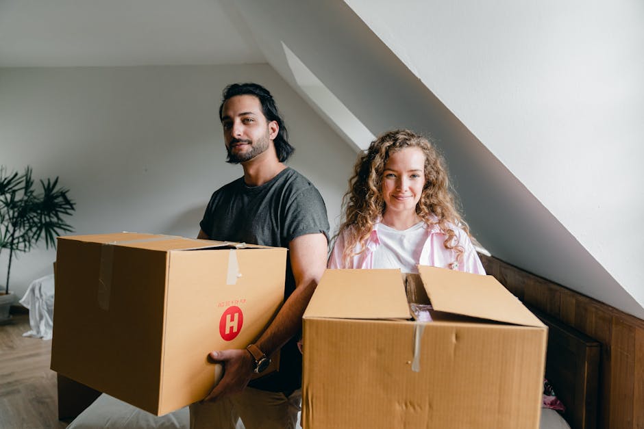 A man with dark hair tied back and a short beard, wearing a dark grey t-shirt, stands inside a home near a sloped ceiling, holding a large cardboard box with a red 'H' symbol on the side. Beside him,