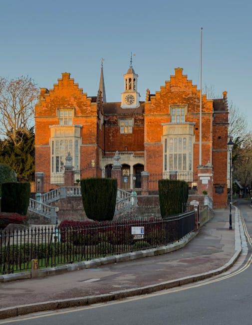 A large historic red brick building with gothic architectural features, including stepped gables, tall narrow windows with stone surrounds, and a central clock tower with a weather vane on top, situated on a well-maintained street. The building is surrounded by a black wrought iron fence with decorative elements, enclosing a landscaped garden with trimmed bushes and paved pathways. A set of stone steps leads up to the entrance, which is accessed via a small ramp for accessibility. The area appears to be part of a residential or institutional complex, with a paved sidewalk running alongside the building and a street lamp positioned on the pavement nearby. The scene is captured in daylight with a clear sky, highlighting the building’s historic architectural details, which may relate to the heritage of the local area and potential considerations during house removals or relocation logistics involving their historic structures. Man with Van West Harrow’s services often include careful planning for loading and transporting such substantial and architecturally significant properties.