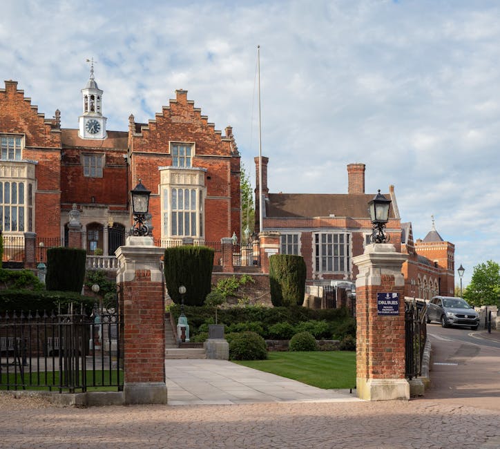 A photograph of a large, historic red brick building with ornate architectural details, including tall, multi-pane windows and a clock tower with a weather vane on top, situated behind a gated entrance. In the foreground, there is a paved pathway leading to the property, flanked by brick pillars with decorative black iron gates, two classic-style black lamp posts, and neatly trimmed bushes and small trees. The setting appears to be in a well-maintained estate or institutional grounds, with a clear sky overhead. This scene is representative of a substantial house or estate that may be involved in home relocation or moving services, as provided by Man with Van West Harrow, who may assist with furniture transport and packing during house removals.