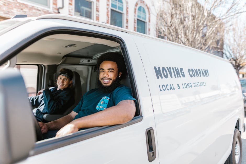 Inside the cab of a white moving van belonging to Man with Van West Harrow, a professional house removals company, two movers are visible. The driver, a man with dark curly hair and a beard, is smiling at the camera and is wearing a blue t-shirt with a small logo. The passenger, a woman with dark hair tied back and a headband, is seated behind him, facing slightly to the side. The van's interior shows part of the dashboard and the driver’s seat, with natural daylight illuminating the scene. The exterior of the van displays the company's name along with the description 'Local & Long Distance'. The vehicle is parked outside a residential property, suggesting the start or end of a home relocation or furniture transport process. This context supports professional packing and moving services, with a focus on safe, efficient loading and transportation of household items.