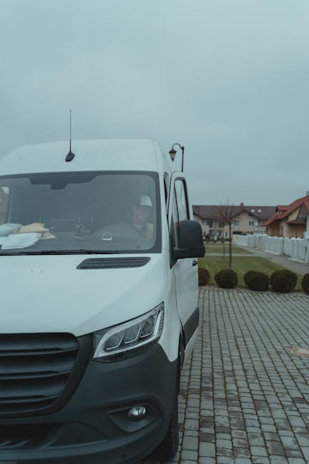 A white van parked on a paved driveway in a residential area, with the front of the vehicle facing slightly to the left and the driver visible through the windshield. The van's driver appears to be preparing for a home relocation or furniture transport, with packing materials such as cardboard boxes and plastic wrap on the dashboard. The van's side mirror is extended, and the vehicle is positioned close to the curb alongside a row of small, well-maintained bushes. In the background, there are suburban houses with red tiled roofs, white fences, and a cloudy sky overhead. The scene demonstrates either the loading or unloading process typical of house removals services provided by Man with Van West Harrow as part of their house moving solutions, supporting efficient packing and logistics for residential relocations in the HA2 area.
