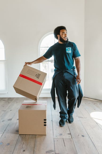 Inside the cab of a white moving van belonging to Man with Van West Harrow, a professional house removals company, two movers are visible. The driver, a man with dark curly hair and a beard, is smiling at the camera and is wearing a blue t-shirt with a small logo. The passenger, a woman with dark hair tied back and a headband, is seated behind him, facing slightly to the side. The van's interior shows part of the dashboard and the driver’s seat, with natural daylight illuminating the scene. The exterior of the van displays the company's name along with the description 'Local & Long Distance'. The vehicle is parked outside a residential property, suggesting the start or end of a home relocation or furniture transport process. This context supports professional packing and moving services, with a focus on safe, efficient loading and transportation of household items.