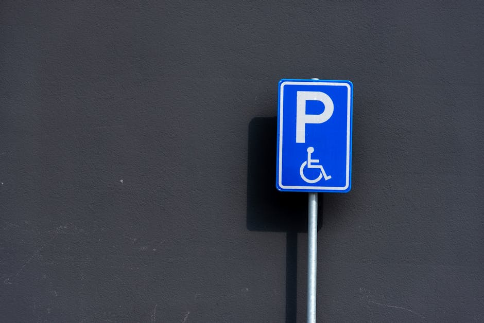 A blue parking permit sign with a white letter 'P' and a wheelchair symbol, mounted on a silver metal pole against a dark, smooth exterior wall. The sign indicates accessible parking, with the shadow of the pole cast on the wall. This image captures the parking permit regulation area relevant to house removals and furniture transport, as part of the moving logistics process. The signage is positioned at the pavement level, outside the property, suggesting a designated space for disabled parking, which Man with Van West Harrow may need to consider during local parking regulations and loading procedures for home relocation services.
