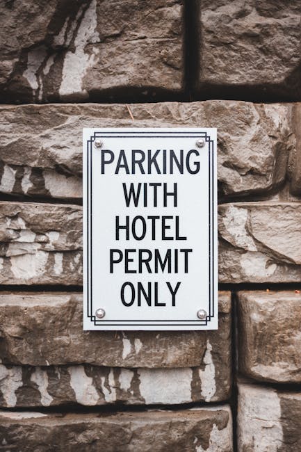 A rectangular metal sign mounted on a stone brick wall, displaying the message 'Parking with hotel permit only' in bold black letters on a white background with a black border. The sign indicates restricted parking regulations, relevant for moves or home relocations in the West Harrow area, as managed by Man with Van West Harrow. The stone wall features irregularly shaped, textured brownish-grey bricks with visible mortar joints. The sign is attached with four screws, one at each corner, and the lighting suggests daytime conditions with natural light illuminating the scene.