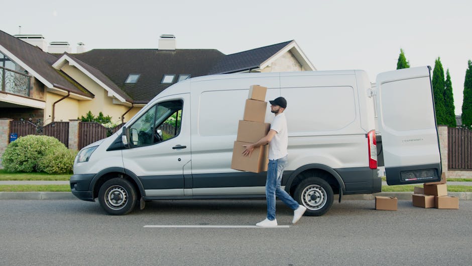 A man wearing a black cap, white t-shirt, and blue jeans is engaged in home relocation activities beside a white moving van parked on a suburban street during daytime. He is carrying three cardboard boxes, one stacked on top of another, with both hands while walking towards the open side door of the van. Additional cardboard boxes are placed on the pavement nearby, ready for loading or unloading. The van is positioned next to a residential property with a modern, two-story house featuring a pitched roof, skylights, and a small front garden with green bushes and a wooden fence. The scene is well-lit with natural daylight, and the vehicle is on the street pavement, indicating an active moving or packing process overseen by a professional removals company, Man with Van West Harrow, specializing in home and furniture transport and packing and moving services.