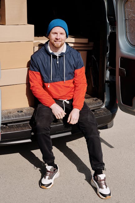 A young man with a beard, wearing a blue beanie hat, a navy and red waterproof jacket, black trousers, and white sneakers, is sitting on the open rear door of a black moving van. Inside the van, several cardboard boxes of varying sizes are stacked, some wrapped in plastic, and wooden furniture pieces are partially visible. The man is smiling and looking at the camera, with his hands resting on his thighs. The van is parked on a paved surface, and the scene is lit by natural daylight. This image depicts a home relocation process involving furniture transport and packing materials, relevant to same-day man with van services provided by Man with Van West Harrow for urgent moves in the Harrow area.