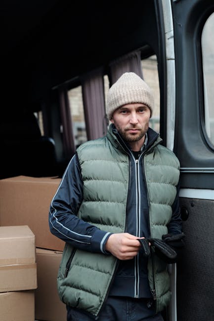 A man wearing a black cap, white t-shirt, and blue jeans is engaged in home relocation activities beside a white moving van parked on a suburban street during daytime. He is carrying three cardboard boxes, one stacked on top of another, with both hands while walking towards the open side door of the van. Additional cardboard boxes are placed on the pavement nearby, ready for loading or unloading. The van is positioned next to a residential property with a modern, two-story house featuring a pitched roof, skylights, and a small front garden with green bushes and a wooden fence. The scene is well-lit with natural daylight, and the vehicle is on the street pavement, indicating an active moving or packing process overseen by a professional removals company, Man with Van West Harrow, specializing in home and furniture transport and packing and moving services.