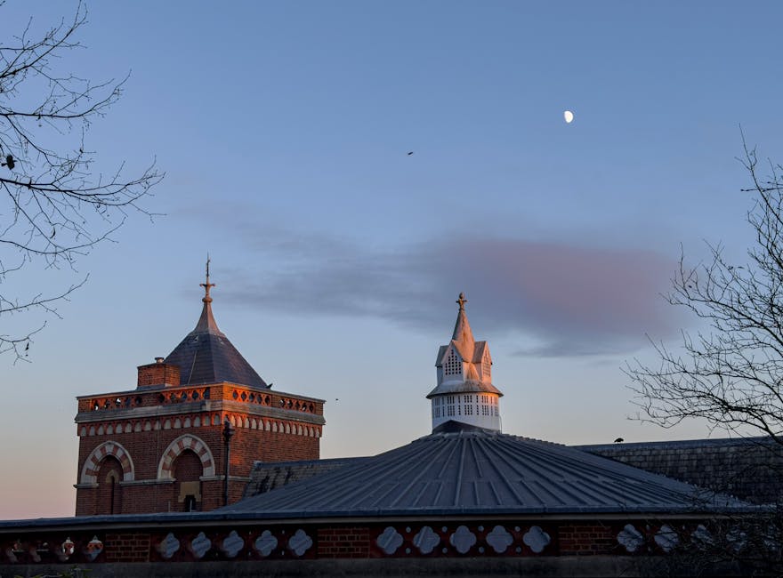 A clear evening sky over a historic brick building with a tall, pointed tower topped with a spire, and an adjacent smaller structure with a pyramidal metal roof. The moon is visible in the night sky, and the branches of leafless trees frame the scene on both sides. This urban landscape is photographed from ground level, capturing the architectural details of the buildings against the twilight sky. As part of house removals or furniture transport, such imagery might be used to illustrate the environment surrounding a residential move near West Harrow, with the structures possibly located close to a station or local landmarks. Man with Van West Harrow ensures careful handling of furniture and belongings during home relocation, with attention to detailed surroundings and external aesthetics essential for smooth logistics and efficient moving processes.