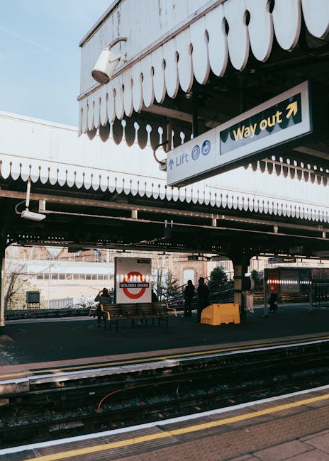 The exterior of West Harrow station showing the platform with black train tracks in the foreground and a yellow safety line along the edge. Above, a metal canopy with scalloped white trim provides shelter, featuring a sign indicating the lift and way out directions. Under the canopy, several individuals are seen standing near a small outdoor seating area with benches and a red London Underground sign displaying 'Golders Green.' To the side, a yellow wheeled trolley is positioned on the pavement, likely used for transporting boxes or furniture during home relocation or moving processes. The station environment is well-lit with natural daylight, emphasizing the functional space where furniture transport and packing are coordinated for quick and efficient house removals, as supported by [COMPANY_NAME] and their services in the [SERVICE_CATEGORY].